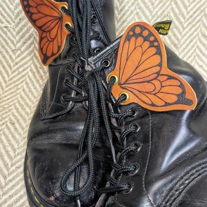 May include: A pair of black leather boots with black laces, adorned with orange butterfly-shaped accessories. The butterfly wings have detailed black outlines. The boots show signs of wear.
