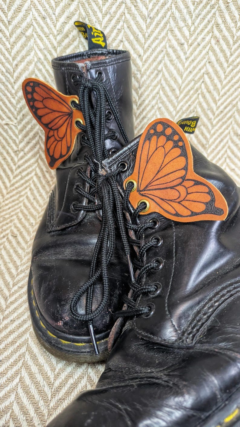 May include: A pair of black leather boots with black laces, adorned with orange butterfly-shaped accessories. The butterfly wings have detailed black outlines. The boots show signs of wear.