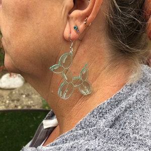 May include: A pair of clear acrylic earrings shaped like a balloon animal dog. The earrings are a light teal color and are hanging from silver hooks.