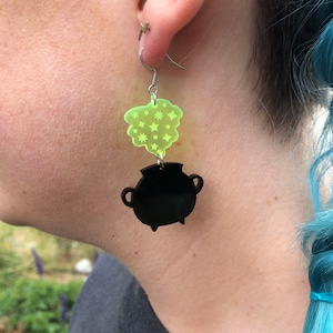 May include: A pair of earrings featuring a black cauldron with green smoke rising from the top. The smoke is decorated with white stars.