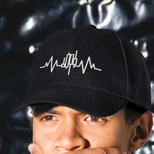 May include: Black baseball cap with a white graphic design resembling a heartbeat. The cap is worn by a person wearing a dark blue sweatshirt. The background is a textured, metallic surface.