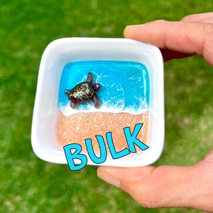 May include: A small white ceramic dish with a resin scene of a sea turtle swimming in blue water with a white foamy wave and a sandy beach. The word "BULK" is written in blue letters on the bottom of the dish.