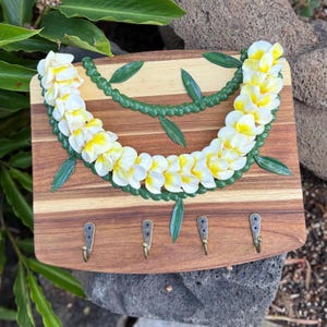 May include: A rectangular wooden wall-mounted key holder featuring a decorative garland of white and yellow plumeria flowers with green leaves. The board has four antique-style metal hooks attached to the bottom section for hanging items.