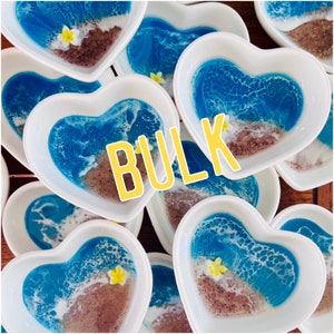 May include: A bulk lot of white heart-shaped ceramic dishes with blue and white resin ocean scene designs. Each dish features a sandy beach, blue waves, and a white flower.