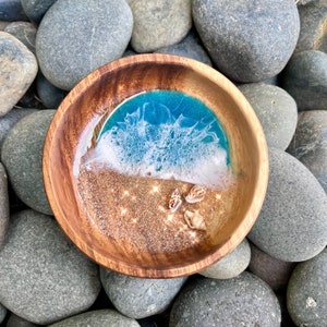 May include: A wooden bowl with a resin ocean scene inside. The resin is blue and white, representing the ocean and waves, with a layer of gold glitter representing sand. There are small seashells in the sand.