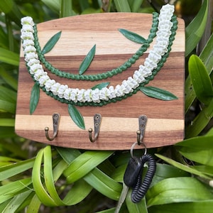 May include: Wooden key holder with three bronze hooks. The holder is decorated with a white and green floral design and green leaf accents. A set of keys hangs from one hook.