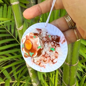 May include: A round, white ornament featuring a miniature food scene. The ornament depicts a fried egg with a bright orange yolk, rice, and a red sauce. Green accents are visible. A white string is attached for hanging.