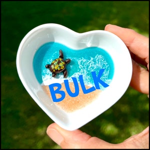 May include: A small, heart-shaped dish with a blue and white resin ocean scene and a tiny turtle figurine. The dish is filled with sand and the word "BULK" is written in blue letters on the sand.