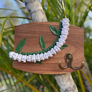 May include: A wooden wall hook with a decorative lei design. The lei is white and green, with green leaves. The hook is made of brass. The wooden board is brown.