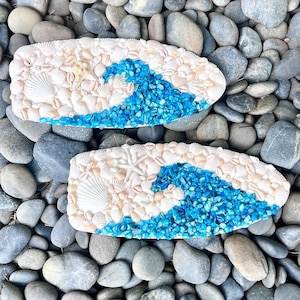 May include: Two rectangular decorative pieces featuring a wave design. The wave is created with blue crushed glass, contrasting with the white and beige seashells and starfish. The pieces are set against a background of gray and brown stones.