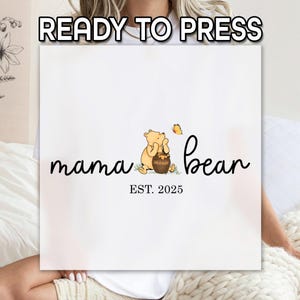 May include: White t-shirt with a black and white graphic of Winnie the Pooh holding a honey pot. The text "mama bear est. 2025" is below the graphic.