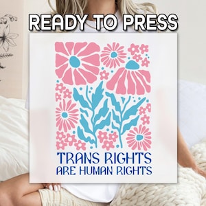 May include: A white square with a floral design in pink and blue, with the words "TRANS RIGHTS ARE HUMAN RIGHTS" in blue. The words "READY TO PRESS" are at the top. The image is on a white background.