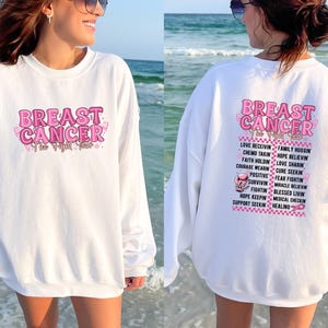 Pu&ograve; includere: Felpa bianca con le parole "BREAST CANCER" in rosa, con un nastro rosa e un design a farfalla. La parte posteriore della felpa presenta un elenco di parole e frasi di supporto in rosa.