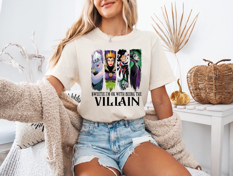 Puede incluir: Camiseta color crema con un dise&ntilde;o de cuatro villanos de dibujos animados: &Uacute;rsula, la Reina Malvada, Cruella de Vil y Mal&eacute;fica. El texto en la camiseta dice: "Sweetie I'm OK with being the Villain".