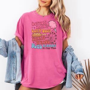 May include: A bright pink t-shirt with a vintage-style design. The text reads "Cotton Candy, Corn Dogs, Ferris Wheel, Blue Ribbons" with graphics of a Ferris wheel and cotton candy. Worn with a light blue denim jacket.