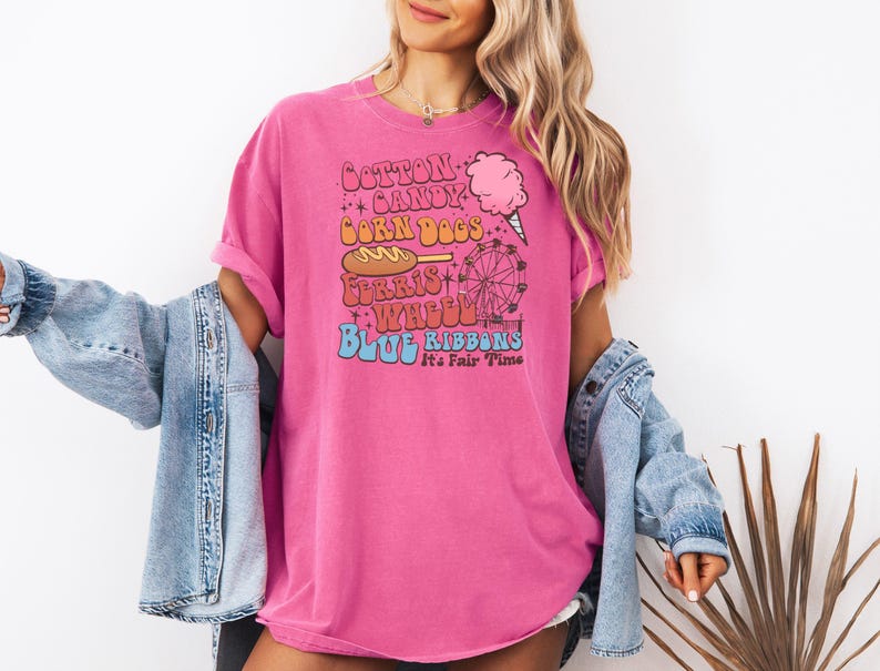 May include: A bright pink t-shirt with a vintage-style design. The text reads "Cotton Candy, Corn Dogs, Ferris Wheel, Blue Ribbons" with graphics of a Ferris wheel and cotton candy. Worn with a light blue denim jacket.