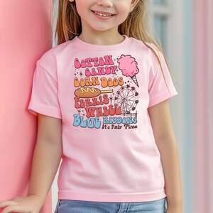 May include: A pale pink t-shirt with the words "Cotton Candy, Corn Dogs, Ferris Wheel, Blue Ribbons" in a retro font. The shirt also has illustrations of a cotton candy cone, a corn dog, and a Ferris wheel. The text "It's Fair Time" is also included.