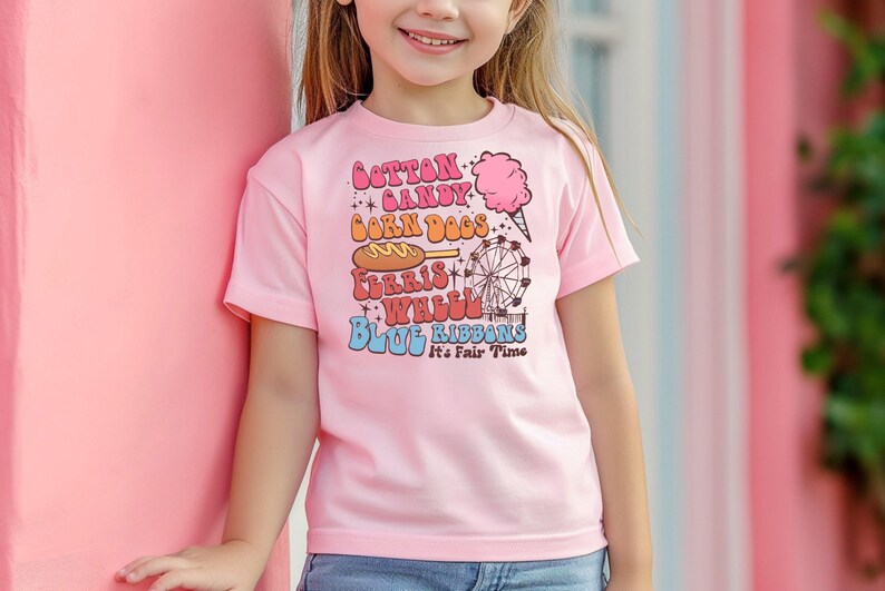 May include: A pale pink t-shirt with the words "Cotton Candy, Corn Dogs, Ferris Wheel, Blue Ribbons" in a retro font. The shirt also has illustrations of a cotton candy cone, a corn dog, and a Ferris wheel. The text "It's Fair Time" is also included.