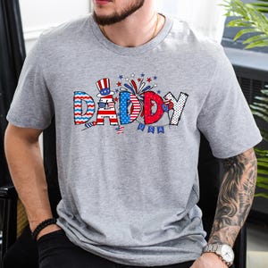 May include: A gray t-shirt with a red, white, and blue graphic that says "Daddy" with fireworks and stars. The graphic is in a cartoon style.