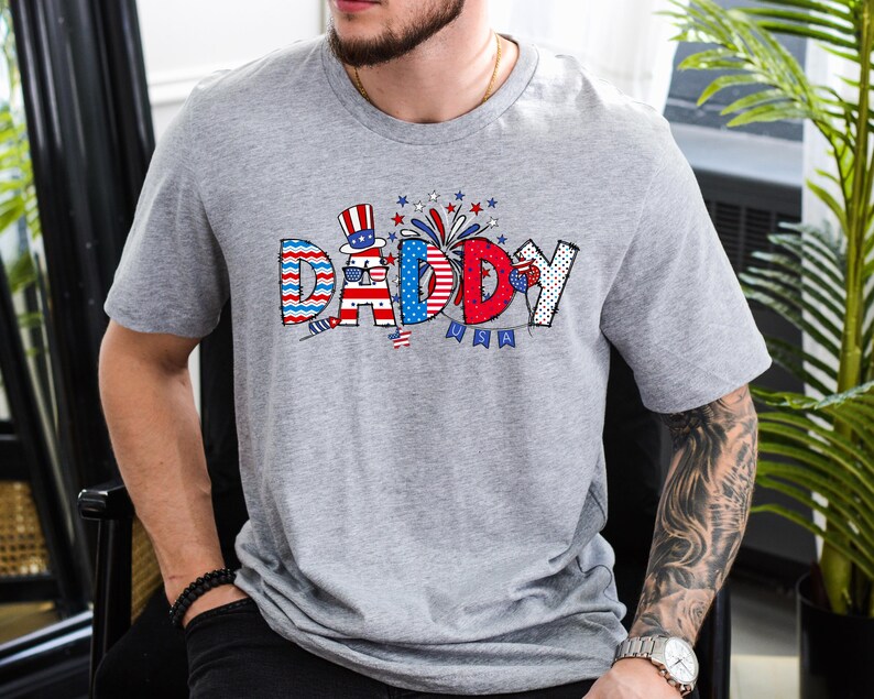 May include: A gray t-shirt with a red, white, and blue graphic that says "Daddy" with fireworks and stars. The graphic is in a cartoon style.