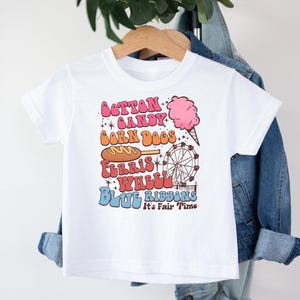 May include: White t-shirt with a colourful graphic featuring carnival-themed text and illustrations. The design includes the words "Cotton Candy," "Corn Dogs," "Ferris Wheel," and "Blue Ribbons," along with images of cotton candy and a Ferris wheel.