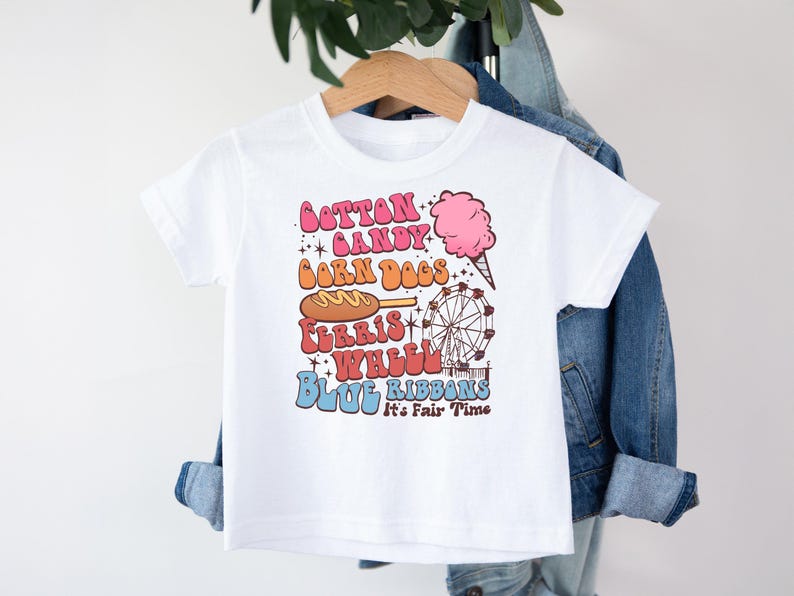 May include: White t-shirt with a colourful graphic featuring carnival-themed text and illustrations. The design includes the words "Cotton Candy," "Corn Dogs," "Ferris Wheel," and "Blue Ribbons," along with images of cotton candy and a Ferris wheel.