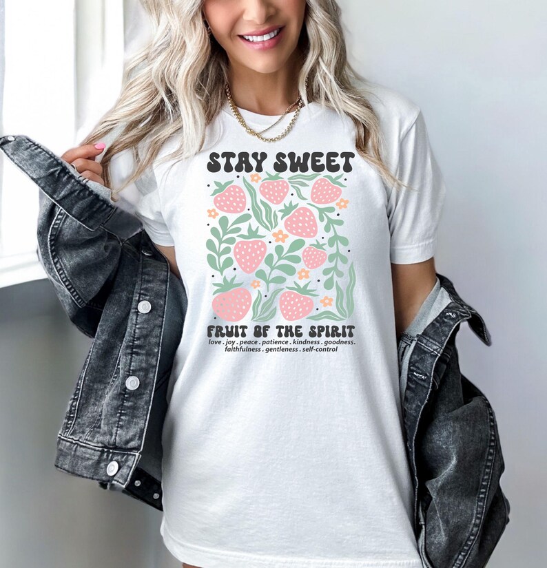 May include: White t-shirt with the text "STAY SWEET" above a graphic of pink strawberries, green leaves, and orange flowers. Below the graphic, the text reads "FRUIT OF THE SPIRIT" and lists virtues. The shirt is worn with a denim jacket.