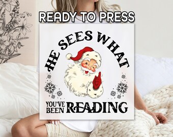 Può includere: Un cartello quadrato bianco con la scritta "READY TO PRESS" in alto. Il cartello presenta un'illustrazione vintage di Babbo Natale con la scritta "HE SEES WHAT YOU'VE BEEN READING" in un design circolare. Sono inclusi anche fiocchi di neve.
