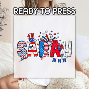 May include: A white t-shirt with a red, white, and blue design featuring the name "SARAH" with a patriotic theme. The design includes stars, stripes, fireworks, and a small American flag.