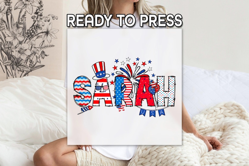 May include: A white t-shirt with a red, white, and blue design featuring the name "SARAH" with a patriotic theme. The design includes stars, stripes, fireworks, and a small American flag.
