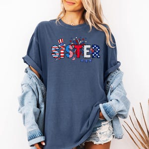 May include: A navy blue t-shirt with a red, white, and blue graphic that says "Sister" with fireworks and a small American flag.