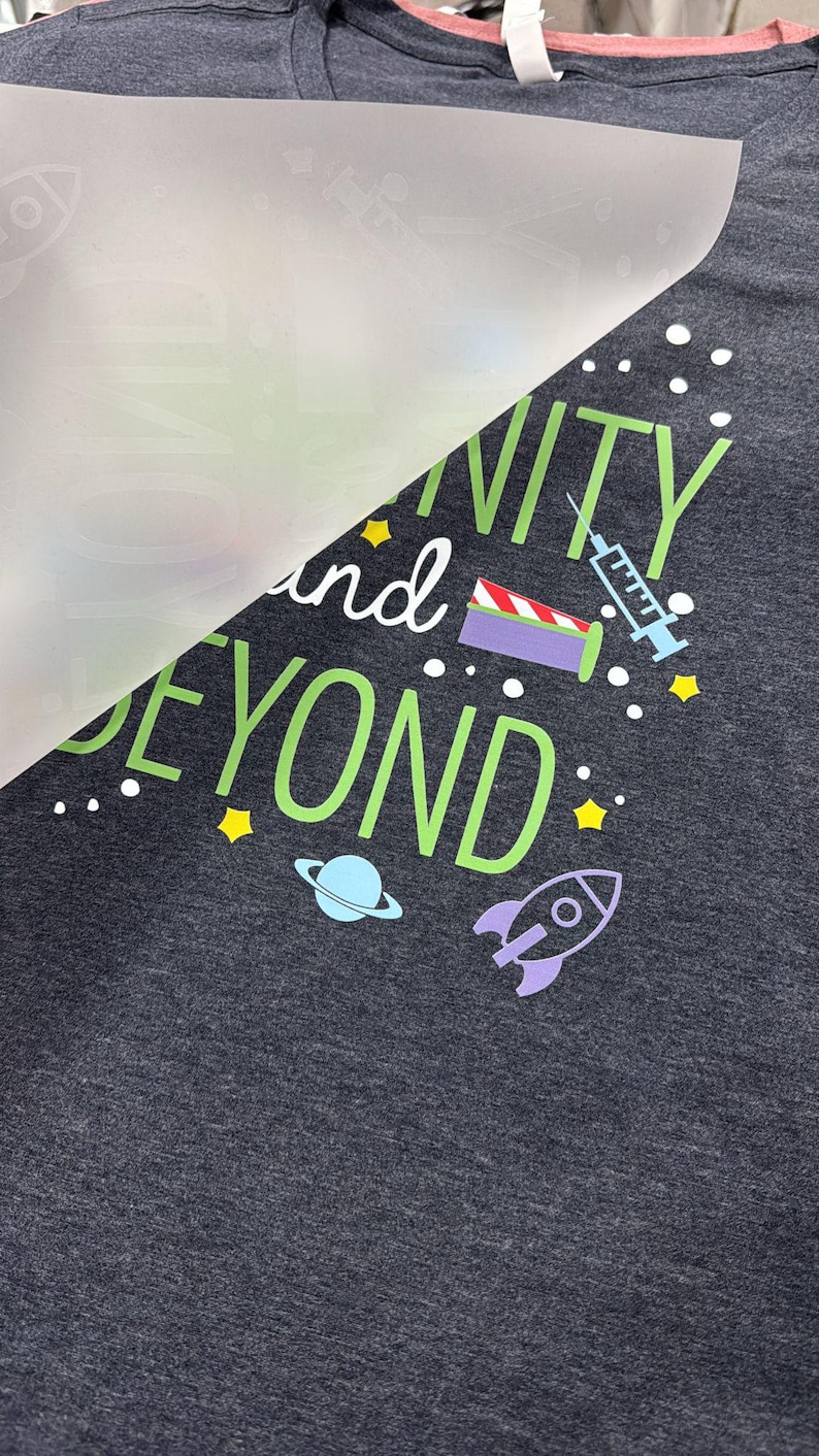 May include: A dark gray t-shirt with a green design that says "Infinity and Beyond" with a rocket, a planet, and a syringe. The design is covered with a clear sheet of paper.