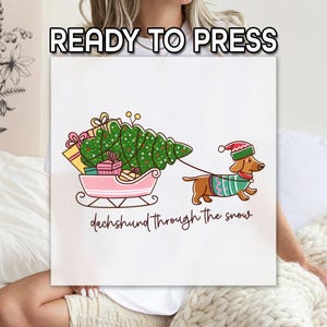 May include: A white canvas with a cartoon illustration of a dachshund pulling a pink sleigh filled with gifts and a Christmas tree. The text "READY TO PRESS" is at the top, and "dachshund through the snow" is written below.