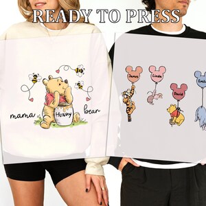May include: Two white sweatshirts with a Winnie the Pooh theme. One sweatshirt has a Winnie the Pooh character with the text "mama bear" and "hunny". The other sweatshirt has a Winnie the Pooh character with the text "James", "Linda", "Emily", and "David".