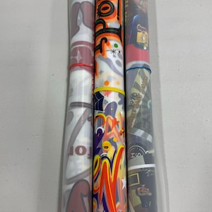 Puede incluir: Tres objetos cil&iacute;ndricos con dise&ntilde;os coloridos. Uno presenta el logotipo de un equipo de f&uacute;tbol, otro tiene la palabra "Hocus Pocus" en naranja y negro, y el tercero muestra un collage de im&aacute;genes. Cada uno est&aacute; asegurado con una goma el&aacute;stica.