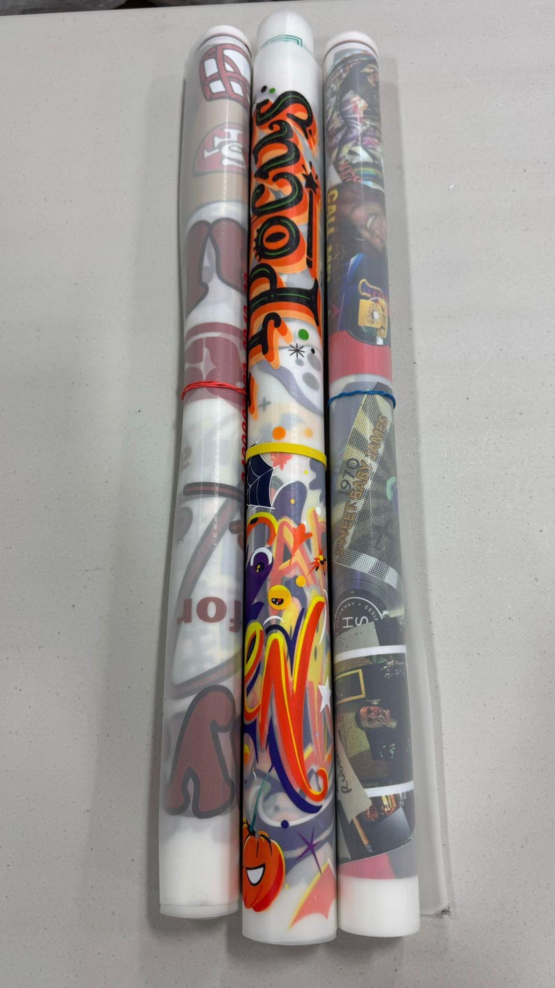 Puede incluir: Tres objetos cil&iacute;ndricos con dise&ntilde;os coloridos. Uno presenta el logotipo de un equipo de f&uacute;tbol, otro tiene la palabra "Hocus Pocus" en naranja y negro, y el tercero muestra un collage de im&aacute;genes. Cada uno est&aacute; asegurado con una goma el&aacute;stica.