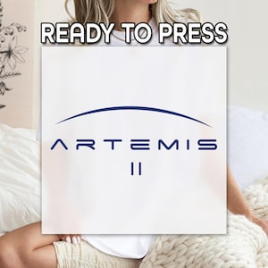 May include: White square with the text "READY TO PRESS" at the top. Below, the word "ARTEMIS" is written in navy blue, with a curved line above it and the number "II" below. The image is held by a person.