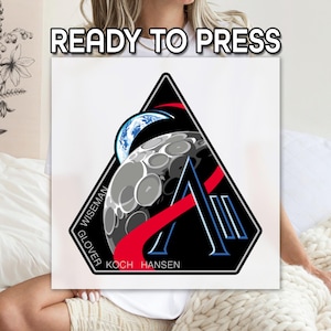May include: A black, white, and red patch with the text "READY TO PRESS" at the top. The patch features a stylized design of the moon, Earth, and a red and white graphic. The names "WISEMAN", "GLOVER", "KOCH", and "HANSEN" are also visible.