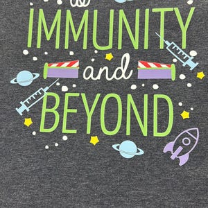 Puede incluir: Camiseta gris oscuro con cuello en V con la frase "to IMMUNITY and BEYOND" en verde y blanco. El dise&ntilde;o incluye planetas, estrellas, jeringas y un cohete. La camiseta es de un material suave.