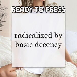 May include: A white square with the text "radicalized by basic decency" is held by a person. Above the square, the text "READY TO PRESS" is displayed. The image is set against a neutral background.