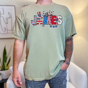 May include: A light green t-shirt with a red, white, and blue graphic of the word "JAMES" with a small American flag and fireworks.