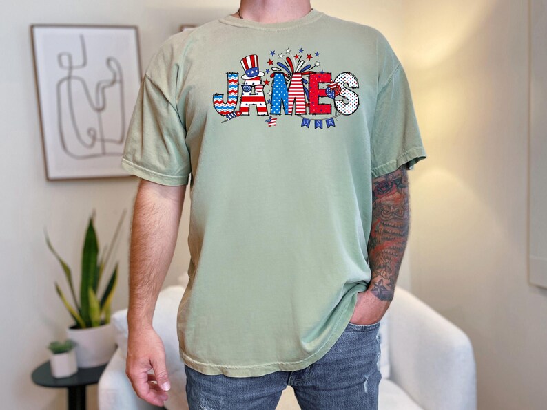 May include: A light green t-shirt with a red, white, and blue graphic of the word "JAMES" with a small American flag and fireworks.