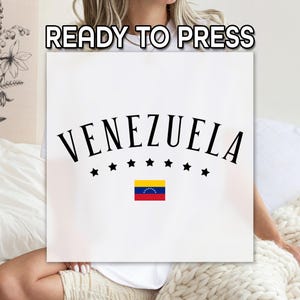 May include: A white square with the word "VENEZUELA" in a curved, black font, with seven black stars below and the Venezuelan flag. The text "READY TO PRESS" is at the top. The image is held by a person.