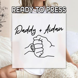 May include: White square with the text "READY TO PRESS" at the top. Below, the words "Daddy + Aidan" are written in a script font. A black line drawing depicts a small hand clasped in a larger hand.