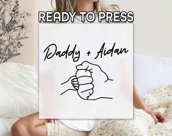 Può includere: Quadrato bianco con la scritta "READY TO PRESS" in alto. Sotto, le parole "Daddy + Aidan" sono scritte in un carattere script. Un disegno a tratto nero raffigura una manina stretta in una mano più grande.