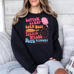 May include: Black sweatshirt with a colourful graphic. The design includes the words "Cotton Candy, Corn Dogs, Ferris Wheel, Blue Ribbons" and a pink cotton candy illustration. The text "It's Fair Time" is also included.