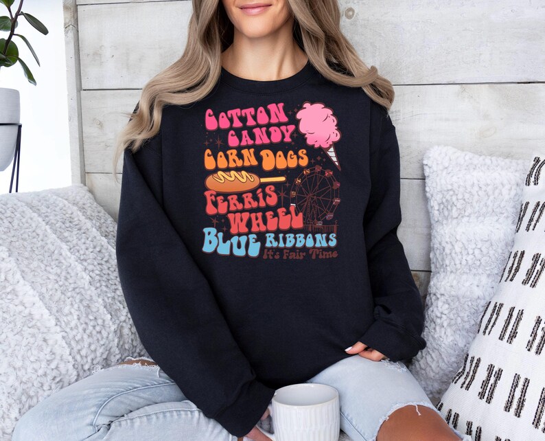 May include: Black sweatshirt with a colourful graphic. The design includes the words "Cotton Candy, Corn Dogs, Ferris Wheel, Blue Ribbons" and a pink cotton candy illustration. The text "It's Fair Time" is also included.