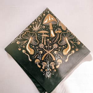 Op de afbeelding: Zwarte bandana met een gouden paddenstoelontwerp. De bandana heeft verschillende paddenstoelen in verschillende vormen en maten, waaronder sommige met witte stippen. Het ontwerp is ingewikkeld en gedetailleerd, en de bandana is perfect om een ​​vleugje whimsy aan elke outfit toe te voegen.