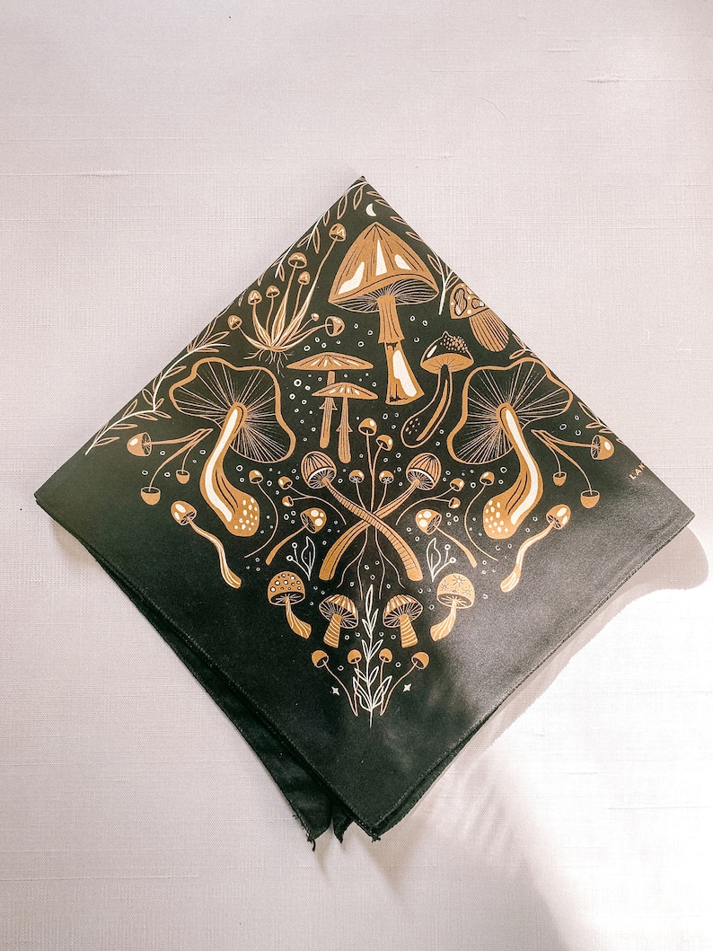 Op de afbeelding: Zwarte bandana met een gouden paddenstoelontwerp. De bandana heeft verschillende paddenstoelen in verschillende vormen en maten, waaronder sommige met witte stippen. Het ontwerp is ingewikkeld en gedetailleerd, en de bandana is perfect om een ​​vleugje whimsy aan elke outfit toe te voegen.