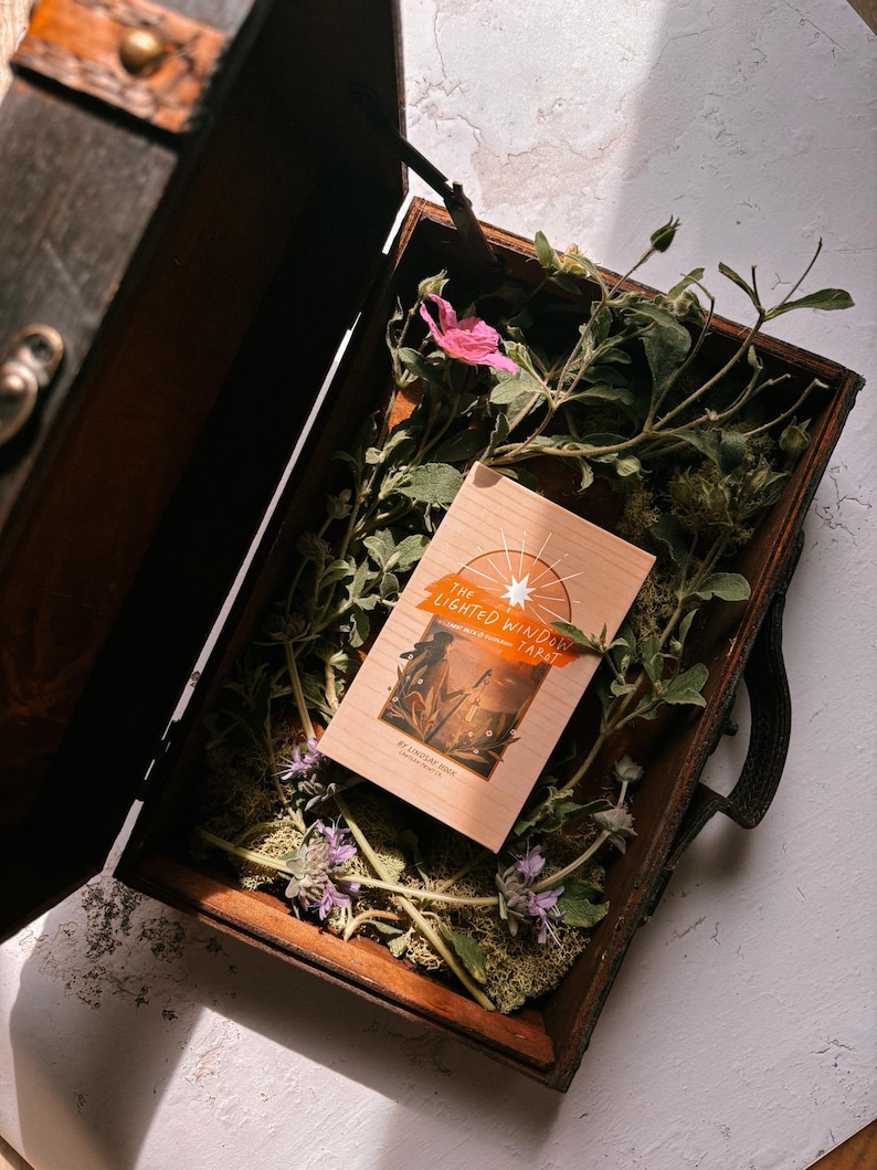 May include: A wooden box, open to reveal a tarot deck titled "The Lighted Window Tarot" surrounded by greenery and flowers. The box is dark brown with metal hardware. The deck has a light brown background with orange and white text.